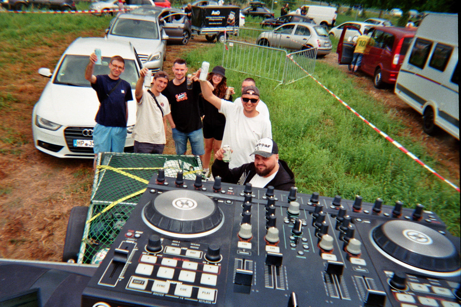 Analoge 35mm-Film-Fotografie einer Gruppe von Freunden, die beim Camping auf dem splash! Festival mit Dosen anstoßen, fotografiert aus der Perspektive hinter einem DJ-Mischpult auf dem Zeltplatz.