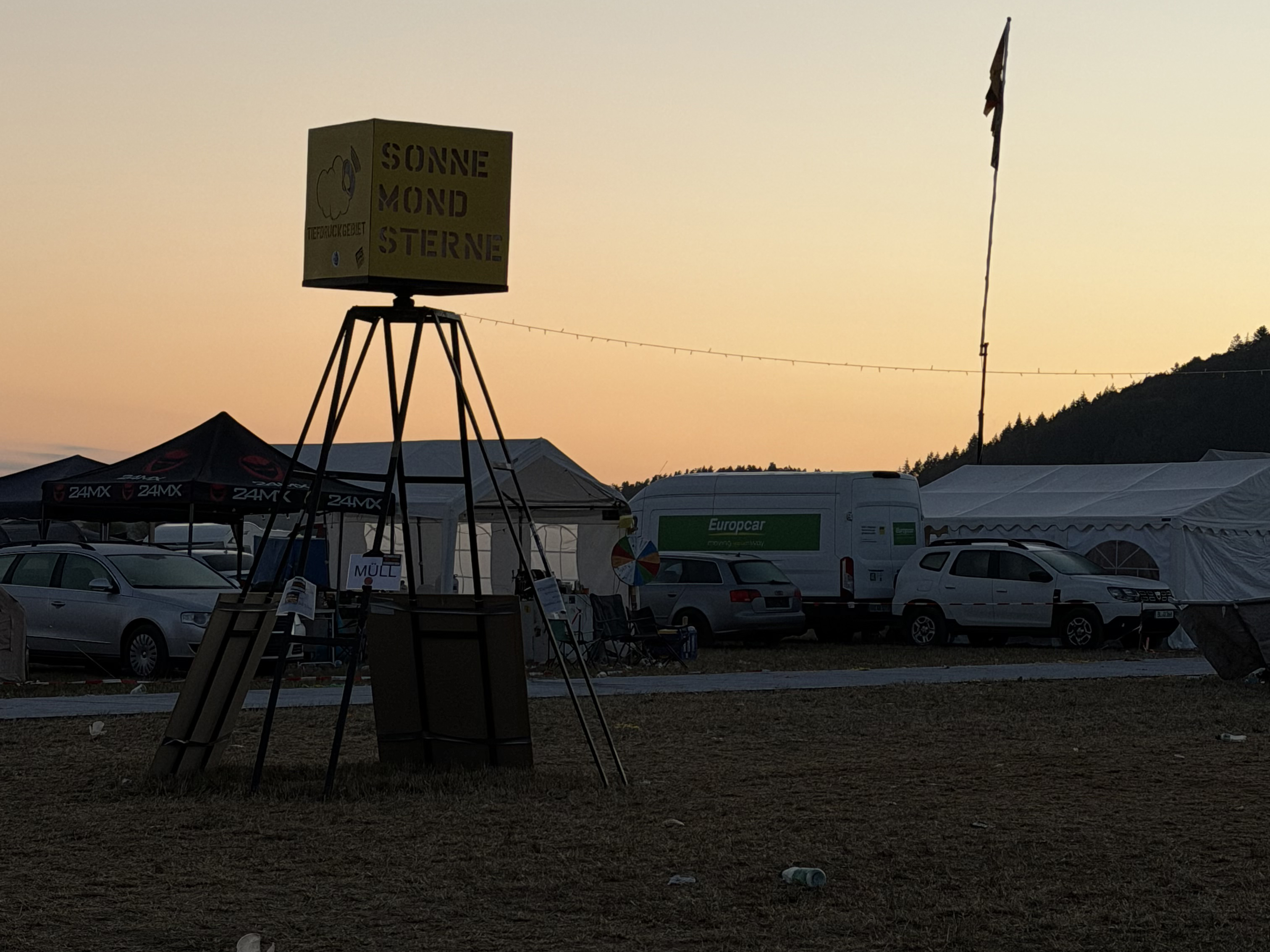 Sonnenaufgang auf dem Campingplatz des SonneMondSterne Festivals. Nach einer langen Festivalnacht taucht die aufgehende Sonne das Gelände mit seinen Zelten, Fahrzeugen und Aufbauten in warmes Morgenlicht. Ein typischer Moment zwischen durchgemachter Partynacht und dem Beginn eines neuen Festivaltages.