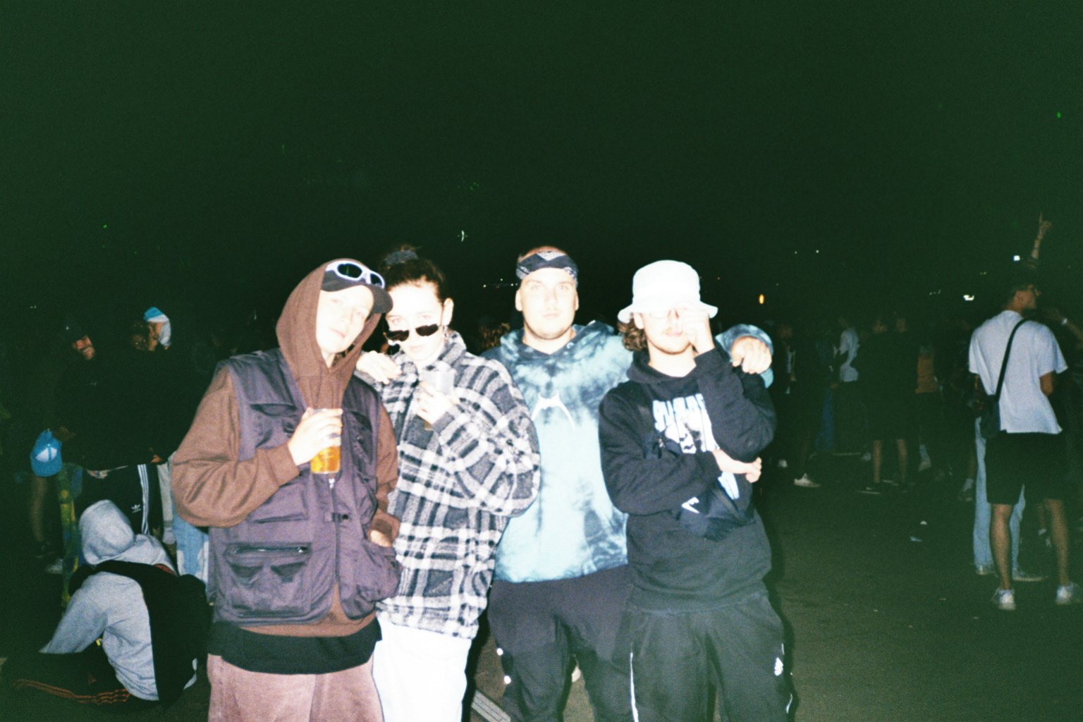 Vier Freunde posieren nachts beim splash! Festival 2022 für ein analoges Gruppenfoto mit Blitzlicht. Sie tragen Hoodies, Sonnenbrillen und einen Bucket Hat, während im Hintergrund weitere Festivalbesucher stehen.