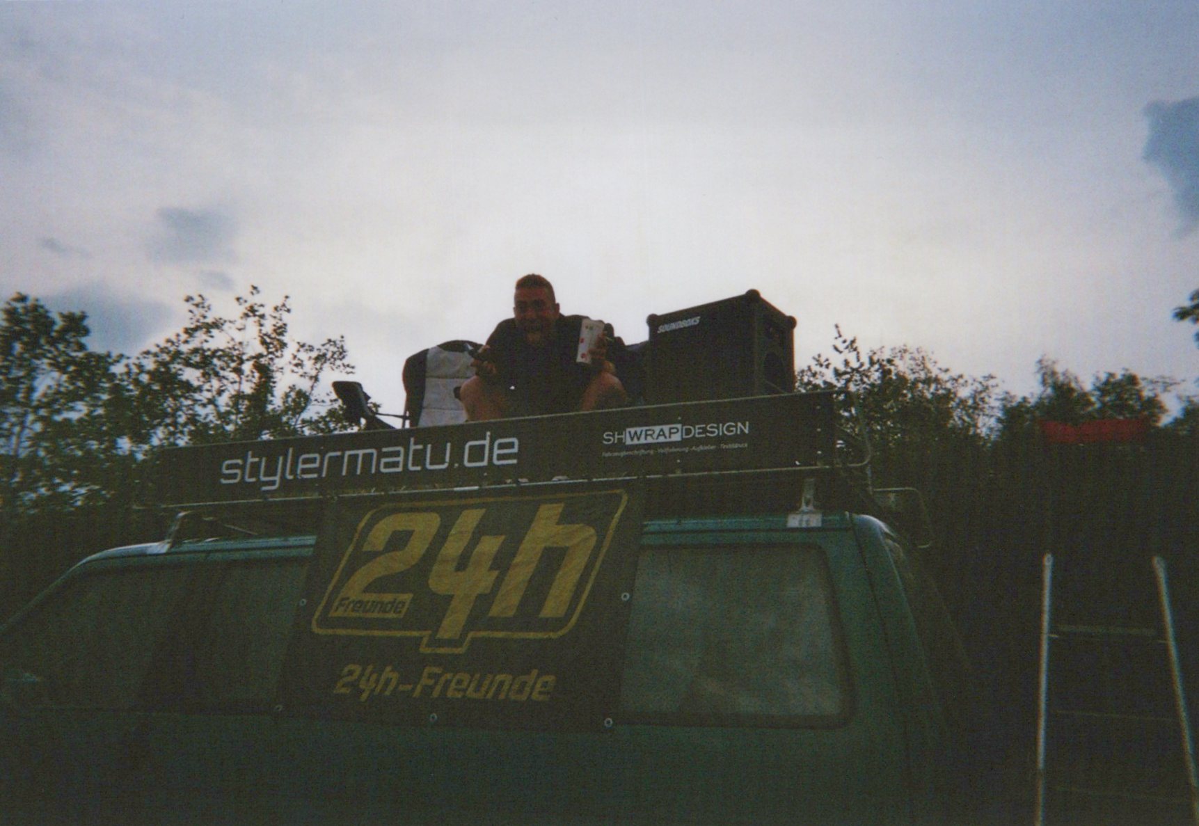 Eine Person sitzt auf dem Dachträger eines Vans auf einem Campingplatz am Nürburgring. Am Fahrzeug sind Banner mit „stylermatu.de“ und „24h-Freunde“ befestigt, daneben steht ein Lautsprecher. Die Aufnahme entstand in der Abenddämmerung während eines Camping- und Motorsport-Wochenendes.