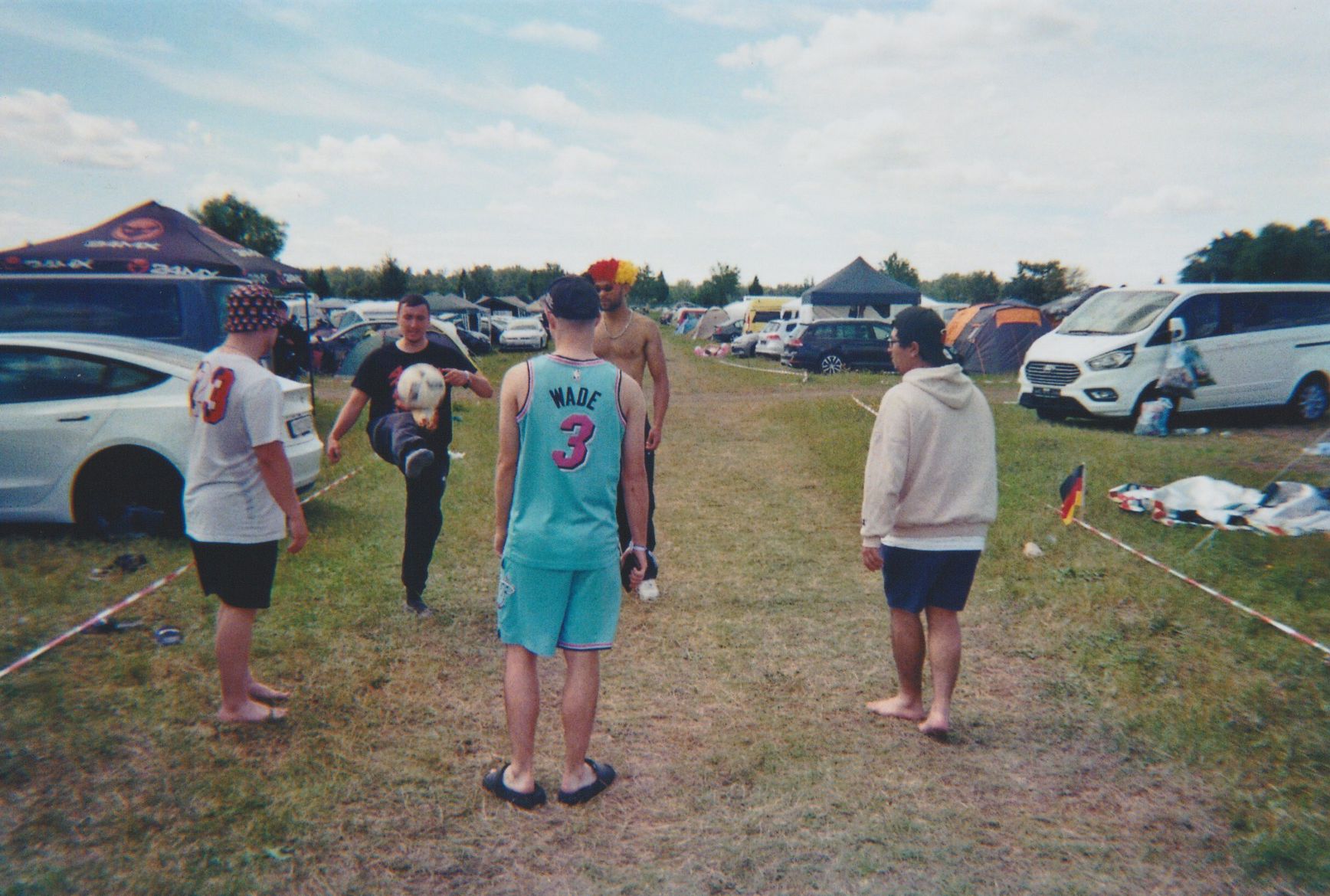 Fünf Personen stehen barfuß oder in Sandalen auf einer Wiese und spielen mit einem Fußball. Sie befinden sich auf einem offenen Gelände mit Zelten und parkenden Autos im Hintergrund. Der Himmel ist hell und leicht bewölkt.