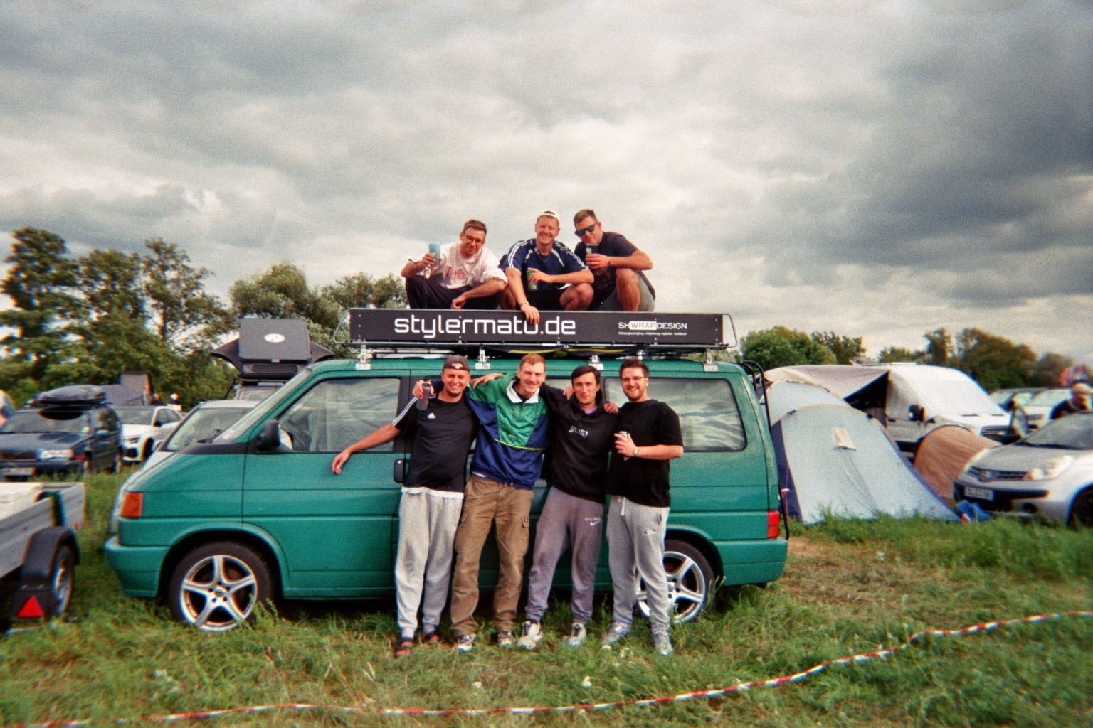 Sieben Freunde posieren beim splash! Festival 2026 vor einem grünen VW-Bus mit stylermatu.de-Banner auf dem Campingplatz.