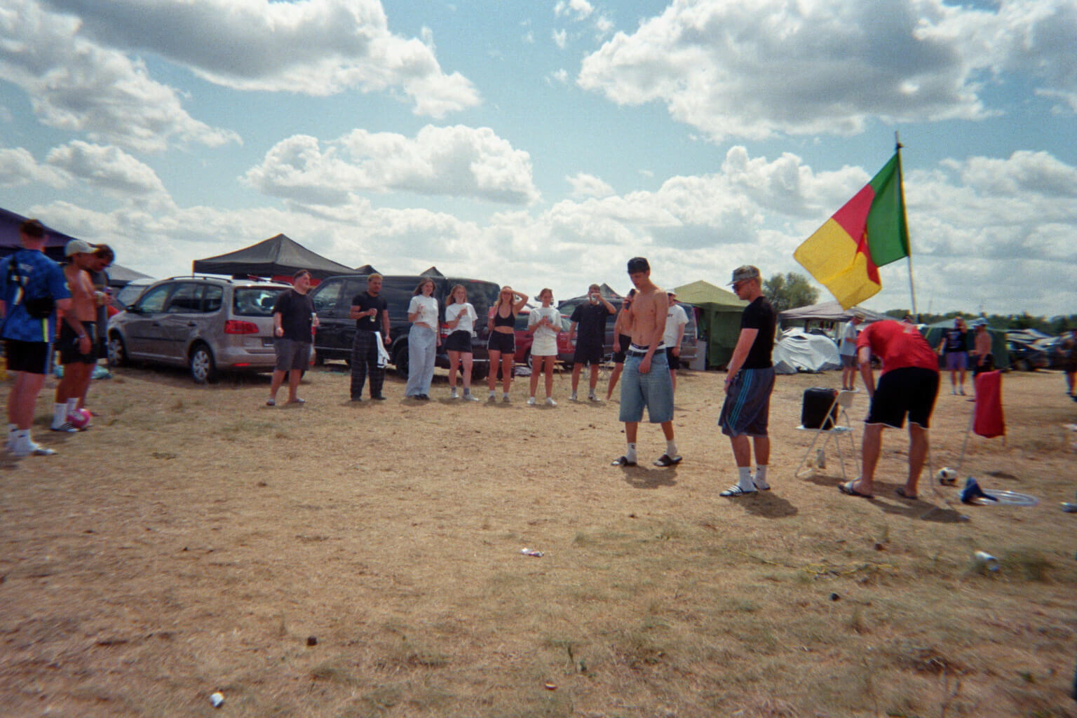 Mehrere Menschen stehen auf einem offenen, staubigen Gelände im Freien. Sie bilden einen Halbkreis um zwei Männer in der Mitte. Im Hintergrund stehen Autos und Zelte. Rechts im Bild weht eine rot-gelb-grüne Flagge, der Himmel ist teilweise bewölkt.