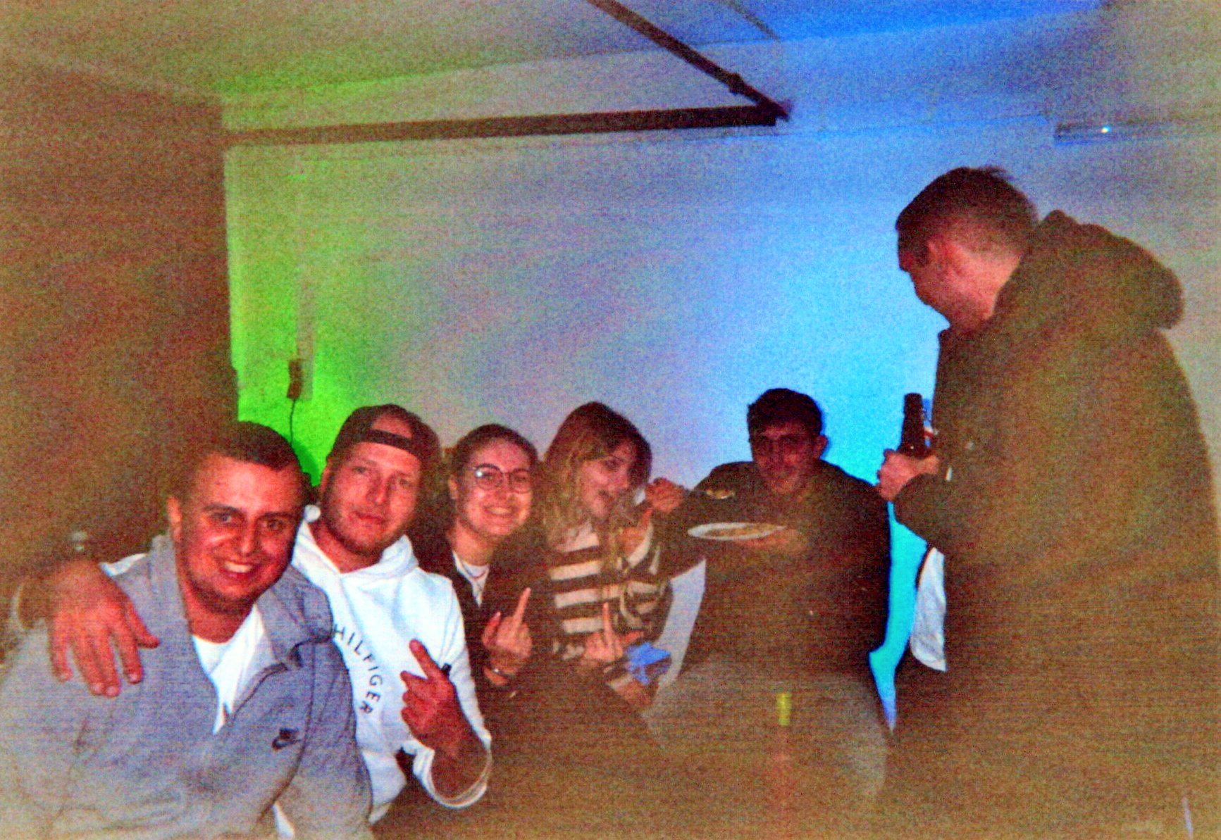 Mehrere Personen sitzen und stehen dicht beieinander in einem Innenraum. Sie lächeln in die Kamera und wirken ausgelassen. Eine Person hält ein Getränk, eine andere einen Teller. Farbiges Licht beleuchtet den Raum im Hintergrund.