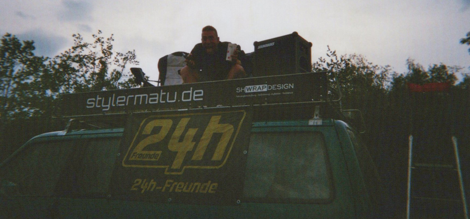 Eine Person sitzt auf dem Dachträger eines Vans auf einem Campingplatz am Nürburgring. Am Fahrzeug sind Banner mit „stylermatu.de“ und „24h-Freunde“ befestigt, daneben steht ein Lautsprecher. Die Aufnahme entstand in der Abenddämmerung während eines Camping- und Motorsport-Wochenendes.