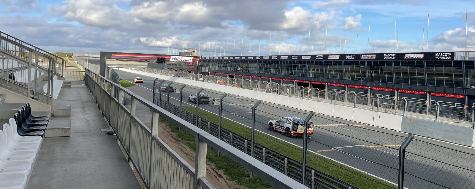 Mehrere Rennwagen fahren auf der Start-Ziel-Geraden des Circuit Zandvoort, rechts die Boxengasse mit Gebäuden, links leere Tribünen unter bewölktem Himmel.
