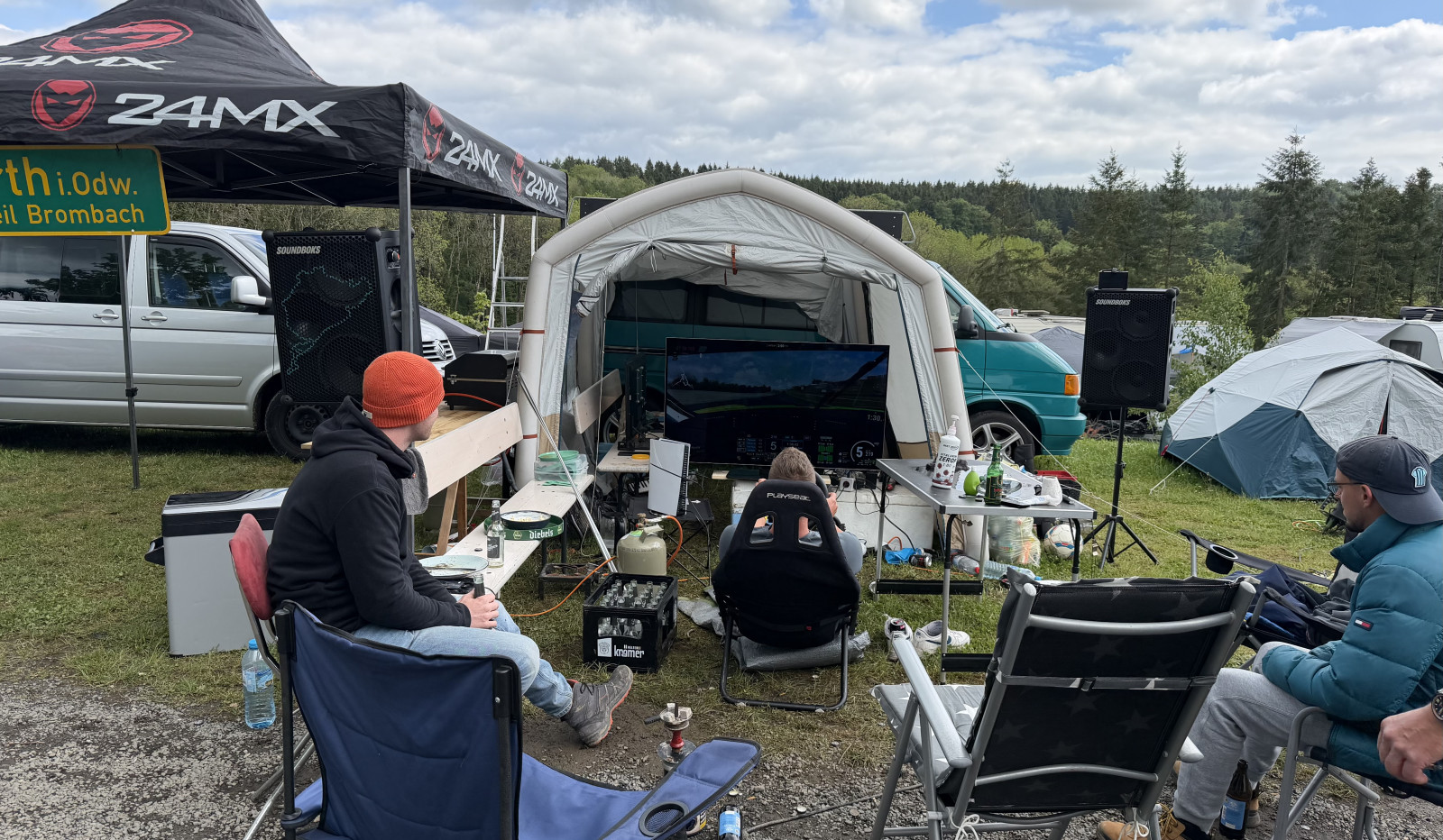 Mehrere Personen sitzen vor einem großen Bildschirm unter einem Pavillon auf einem Campingplatz und spielen gemeinsam ein Rennspiel mit Simracing-Setup. Rundherum stehen Zelte, Transporter und Lautsprecher, im Hintergrund ist ein Wald zu sehen.
