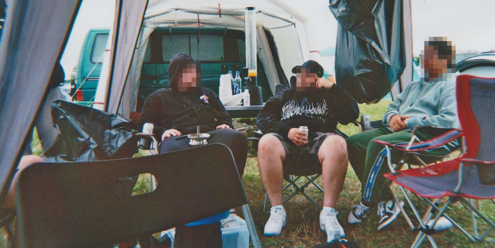 Drei Personen sitzen beim Camping in einem Vorzelt auf Campingstühlen. Auf dem Tisch stehen Getränke und Becher, im Hintergrund ist ein geparkter Transporter zu sehen.