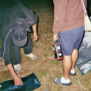 Zwei Personen bauen nachts ein Zelt auf einer Wiese auf, Campingausrüstung liegt auf dem Boden.